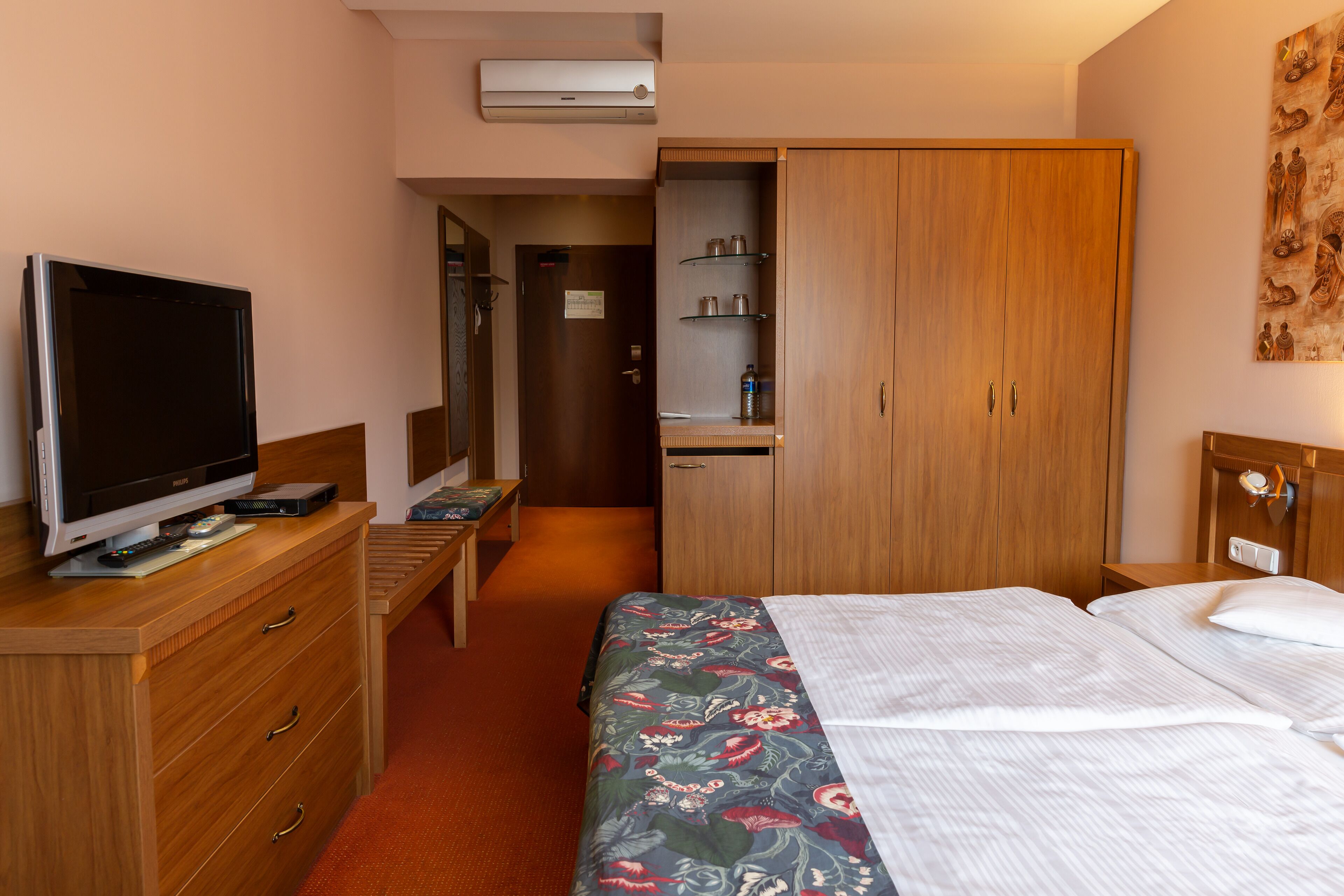 A hotel room with a bed, a TV on a wooden dresser, a wardrobe, and a small desk with a chair.