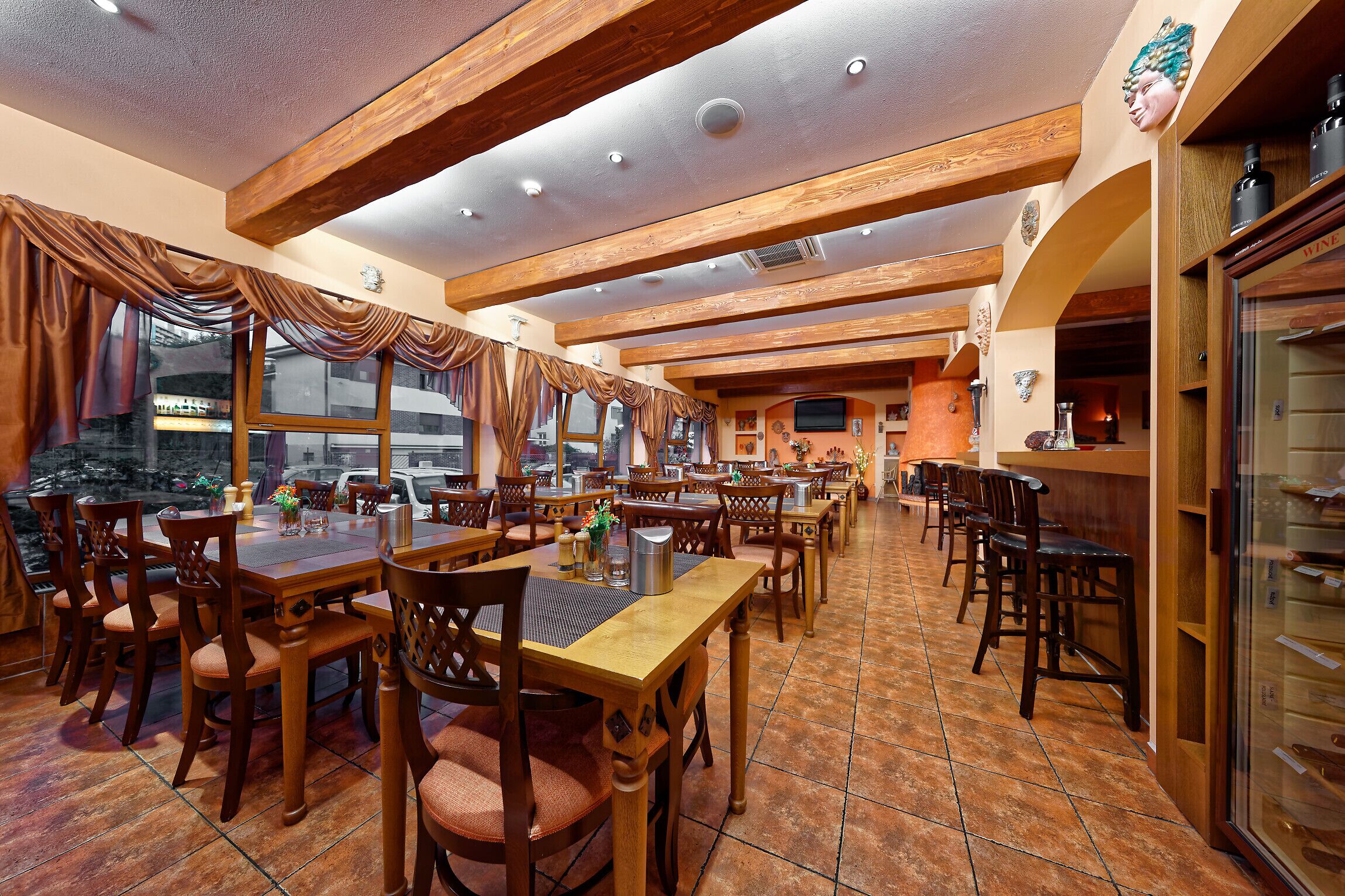 Een restaurant met houten tafels en stoelen, grote ramen met gordijnen en een betegelde vloer.