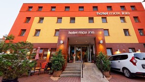 Un hotel llamado 'hotel max inn' con fachada roja y amarilla, una escalera que conduce a la entrada y un coche blanco estacionado al frente.