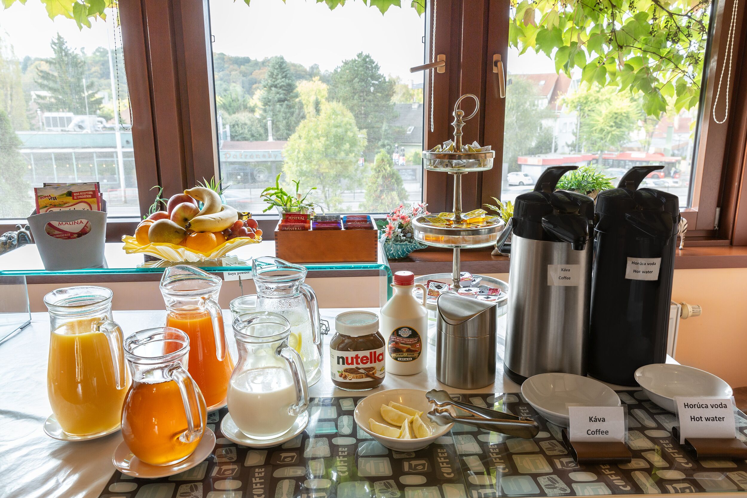 Ein reichhaltiges Frühstücksbuffet mit verschiedenen Speisen und Getränken, darunter Saft, Kaffee und heißes Wasser, präsentiert an einem Tisch in der Nähe eines Fensters mit Blick auf Bäume und Gebäude.