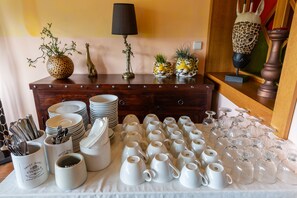 A table with white ceramic mugs and glassware, a wooden cabinet with decorative items, and a lamp. - Max Inn Hotel (Bratislava)