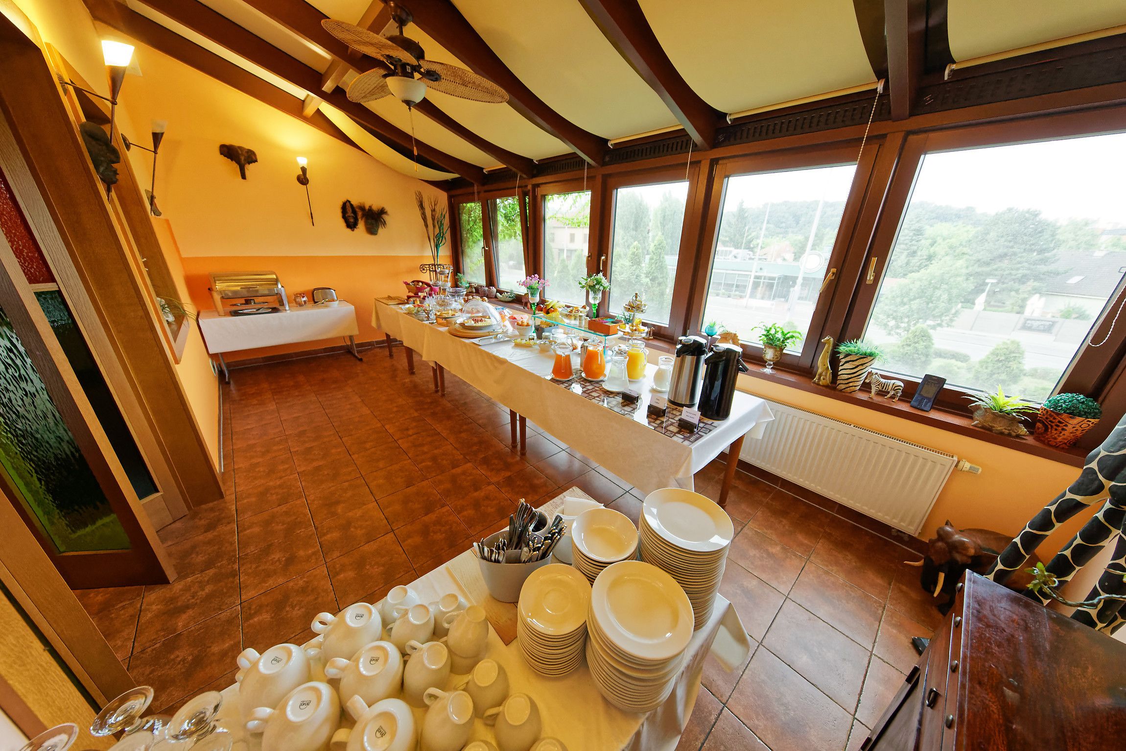 A breakfast buffet setup with various dishes and utensils, a long table, and a view of greenery outside.