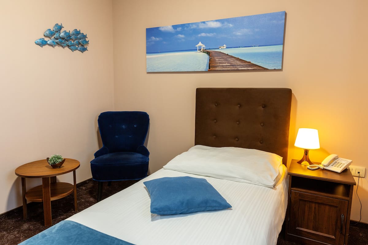 Quarto de hotel com uma cama, uma cadeira azul, uma pequena mesa com uma planta, uma mesa de cabeceira com abajur e um quadro de um cais na parede.