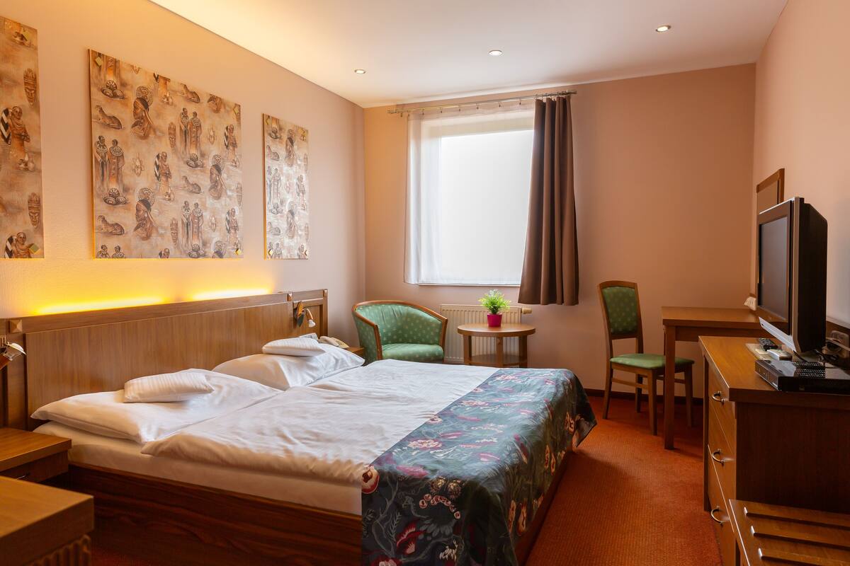 Quarto de hotel com uma cama, uma escrivaninha com um computador, uma cadeira, uma mesinha com uma planta e obras de arte na parede.