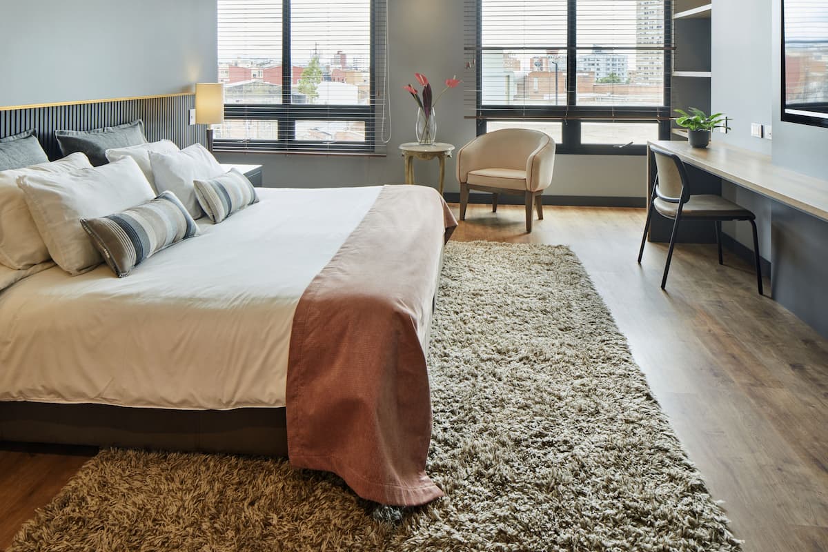 Quarto moderno com uma cama grande, uma mesa de escritório e uma cadeira. Há duas janelas com persianas, vista para edifícios e uma pequena mesa com um vaso de flores.