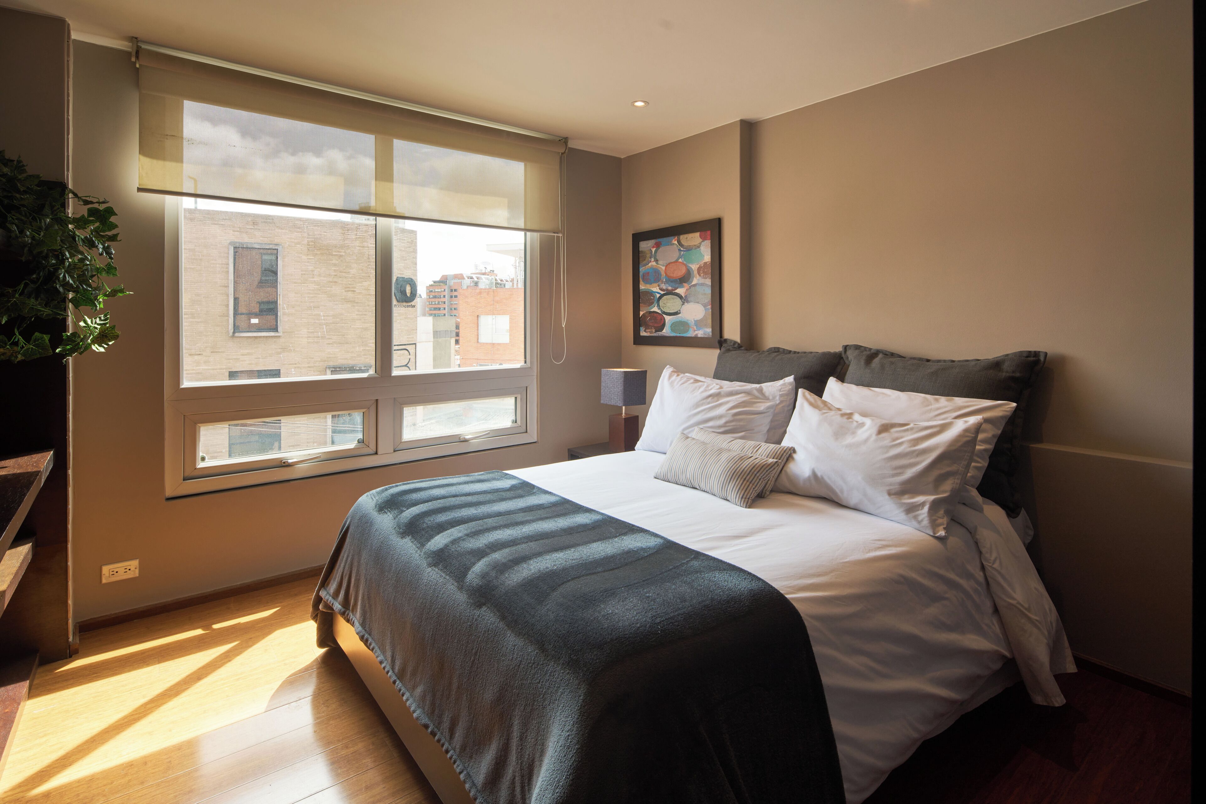 A bedroom with a large window, a bed with white bedding, a grey blanket, and a painting on the wall.