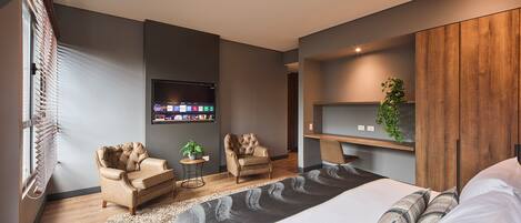 A modern bedroom with a large bed, two leather armchairs, a small table with a plant, a mounted TV, and a wooden shelf with a plant.