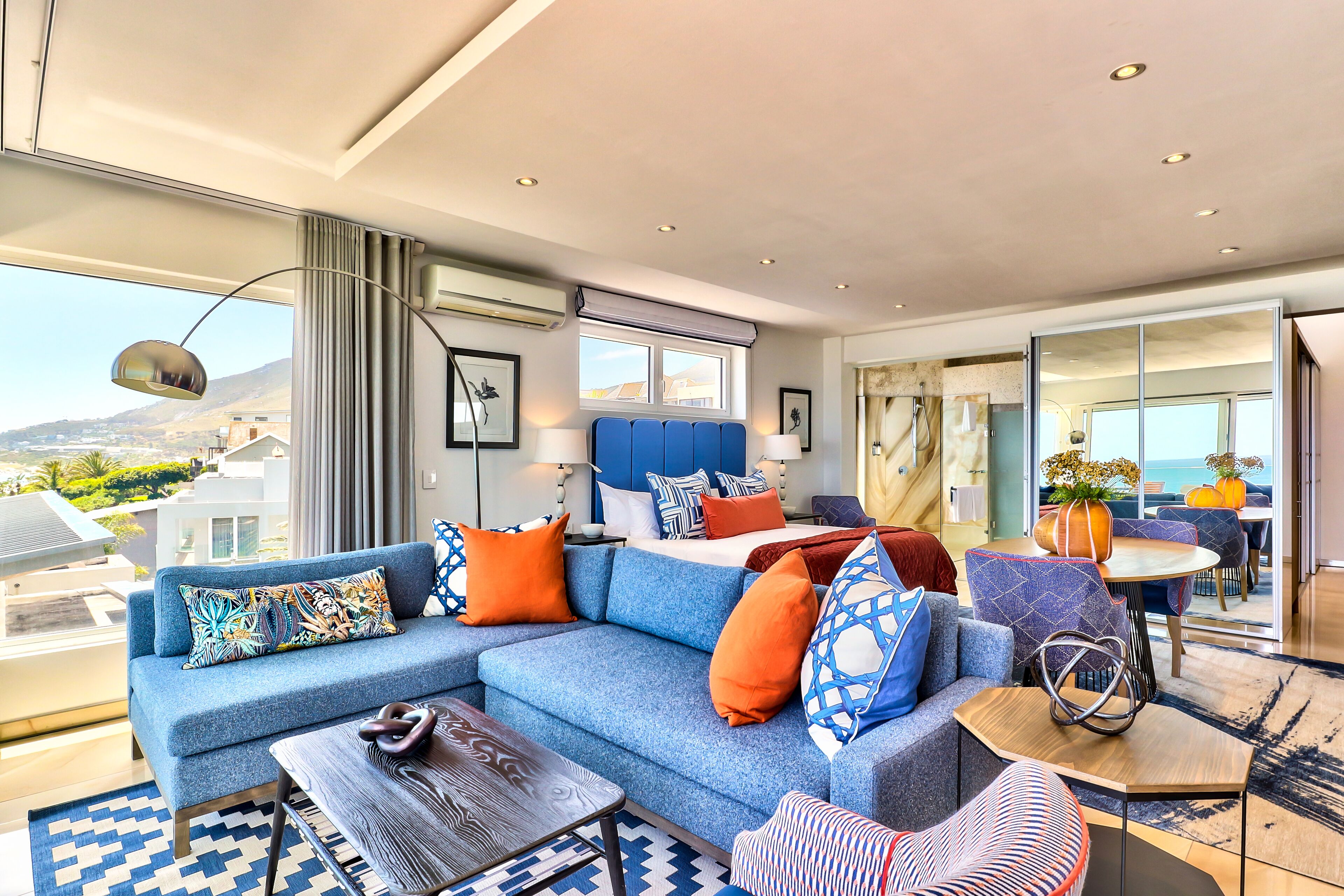 A modern hotel room with a large sofa, a dining table, and a view of the city.