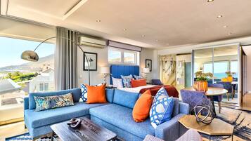A modern hotel room with a large sofa, a dining table, and a view of the city.
