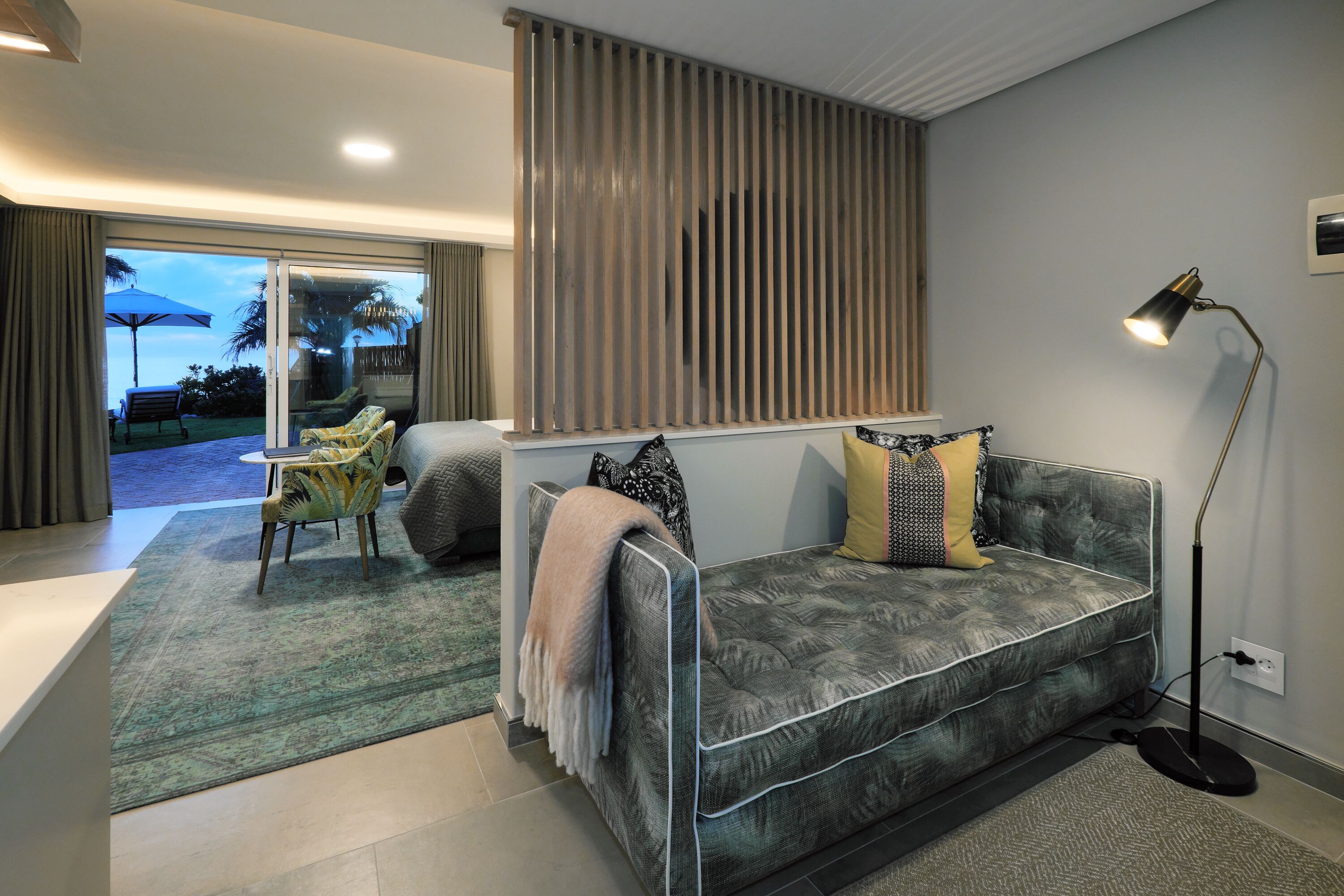 A modern living room with a grey sofa, patterned cushions, a floor lamp, and a view through a sliding glass door to an outdoor area with a table and chairs.