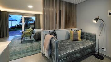 A modern living room with a grey sofa, patterned cushions, a floor lamp, and a view through a sliding glass door to an outdoor area with a table and chairs.