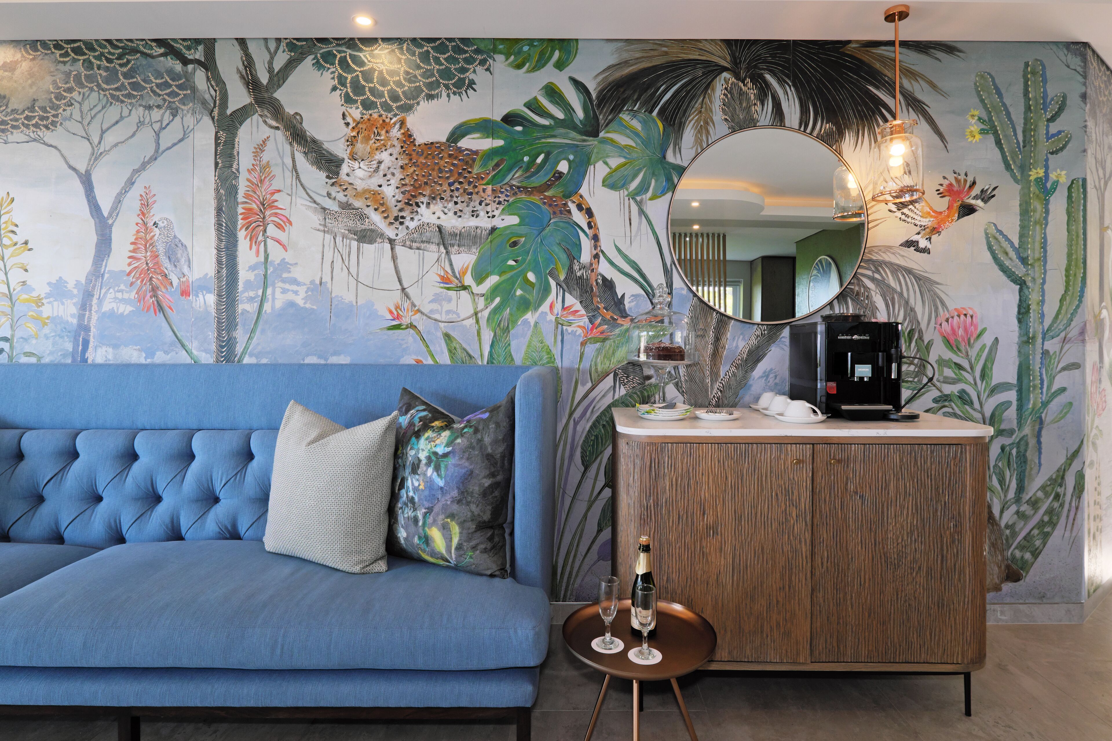 Una habitación con un sofá azul, un aparador de madera, un espejo y un mural con motivos de jungla.