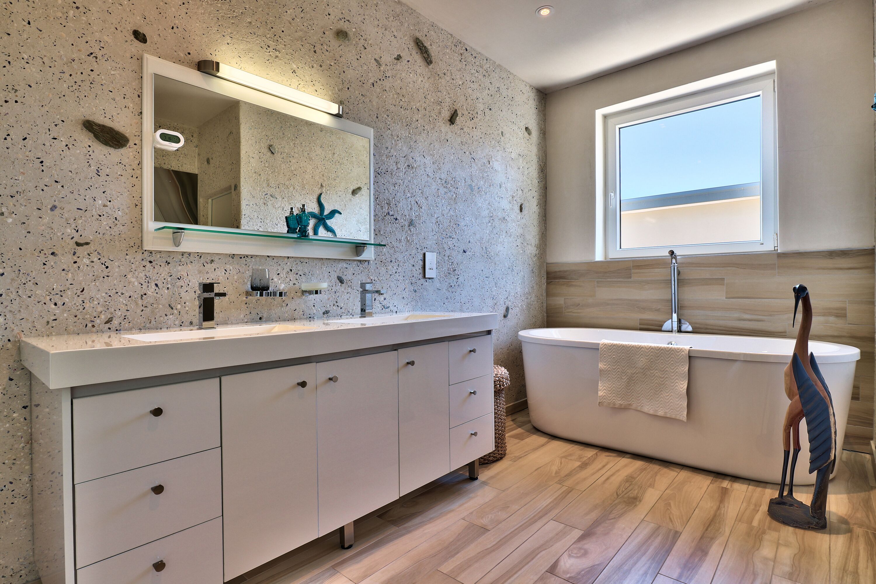 A modern bathroom with a granite speckled wall, a large mirror, a white vanity, a freestanding bathtub, and a window with a view.
