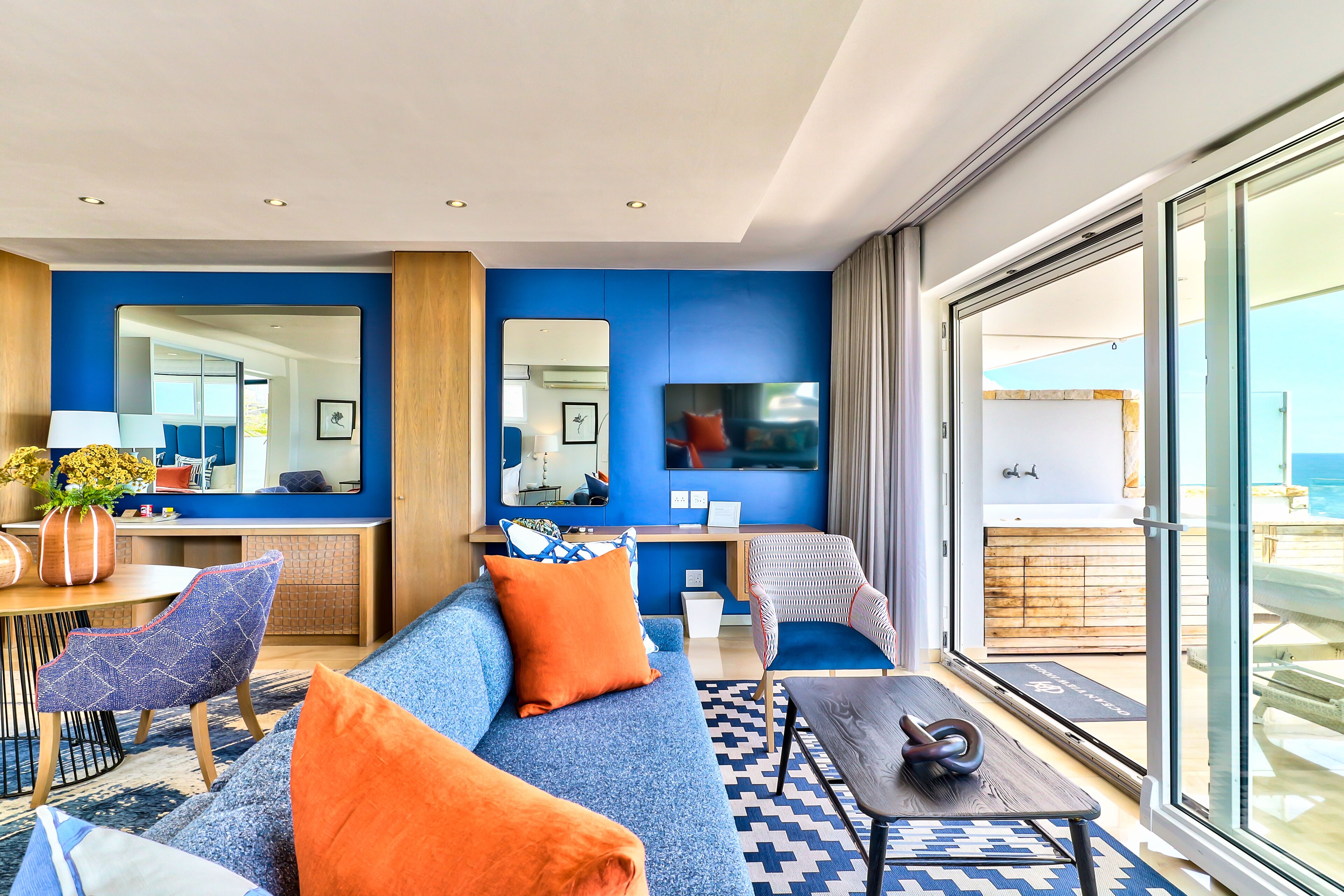 Una habitación de hotel moderna con una pared azul de acento, un sofá con cojines naranjas, una zona de comedor con mesa y sillas, y un balcón con vistas al mar.