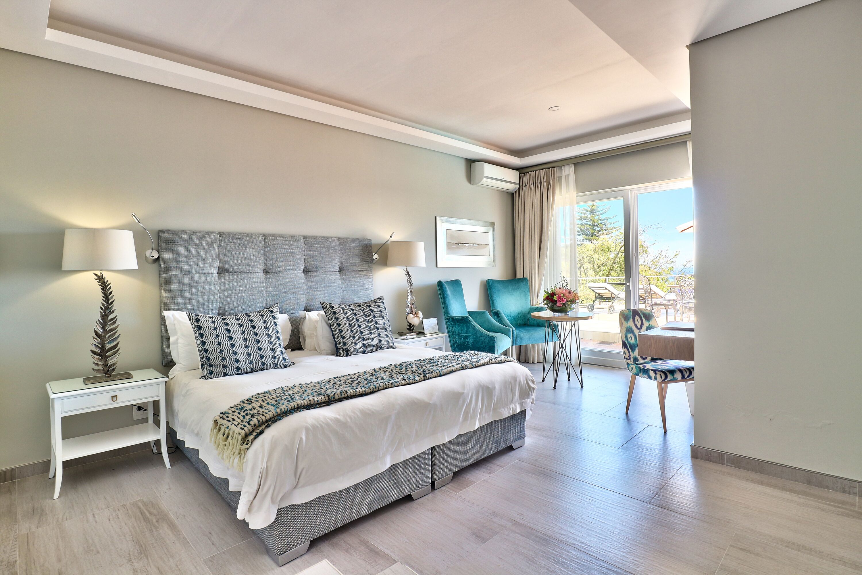 Quarto de hotel moderno com uma cama grande, uma mesa de cabeceira, uma cadeira e uma pequena mesa com um vaso de flores.