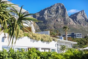 A modern building with a white facade, surrounded by lush greenery and palm trees. In the background, there are two prominent mountains. - Ocean View House (Cape Town)