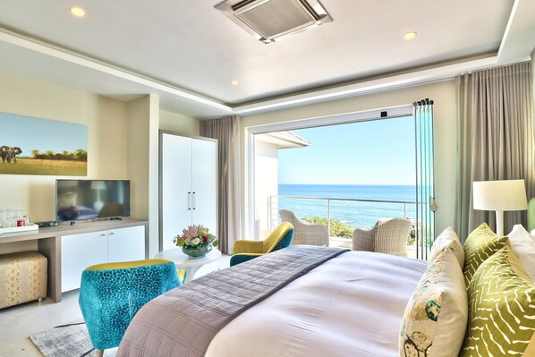 A modern hotel room with a large bed, a desk with a TV, a balcony with a view of the ocean, and a dining area.