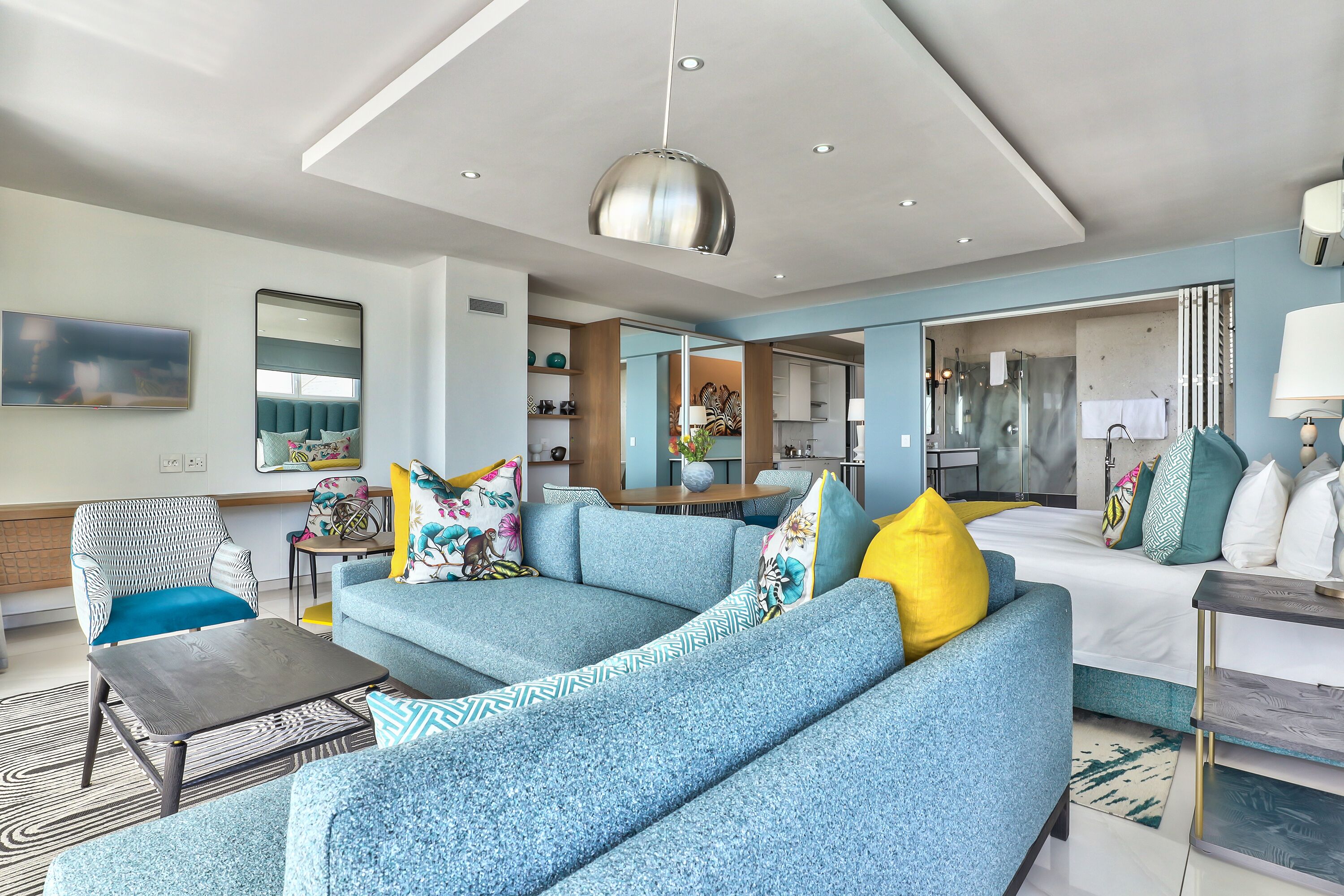 A modern hotel room with a blue sofa, a wooden coffee table, a large mirror, and a hanging pendant light.