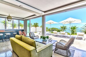 A modern lounge area with a sofa, chairs, and a table. Large windows offer a view of the ocean and palm trees.