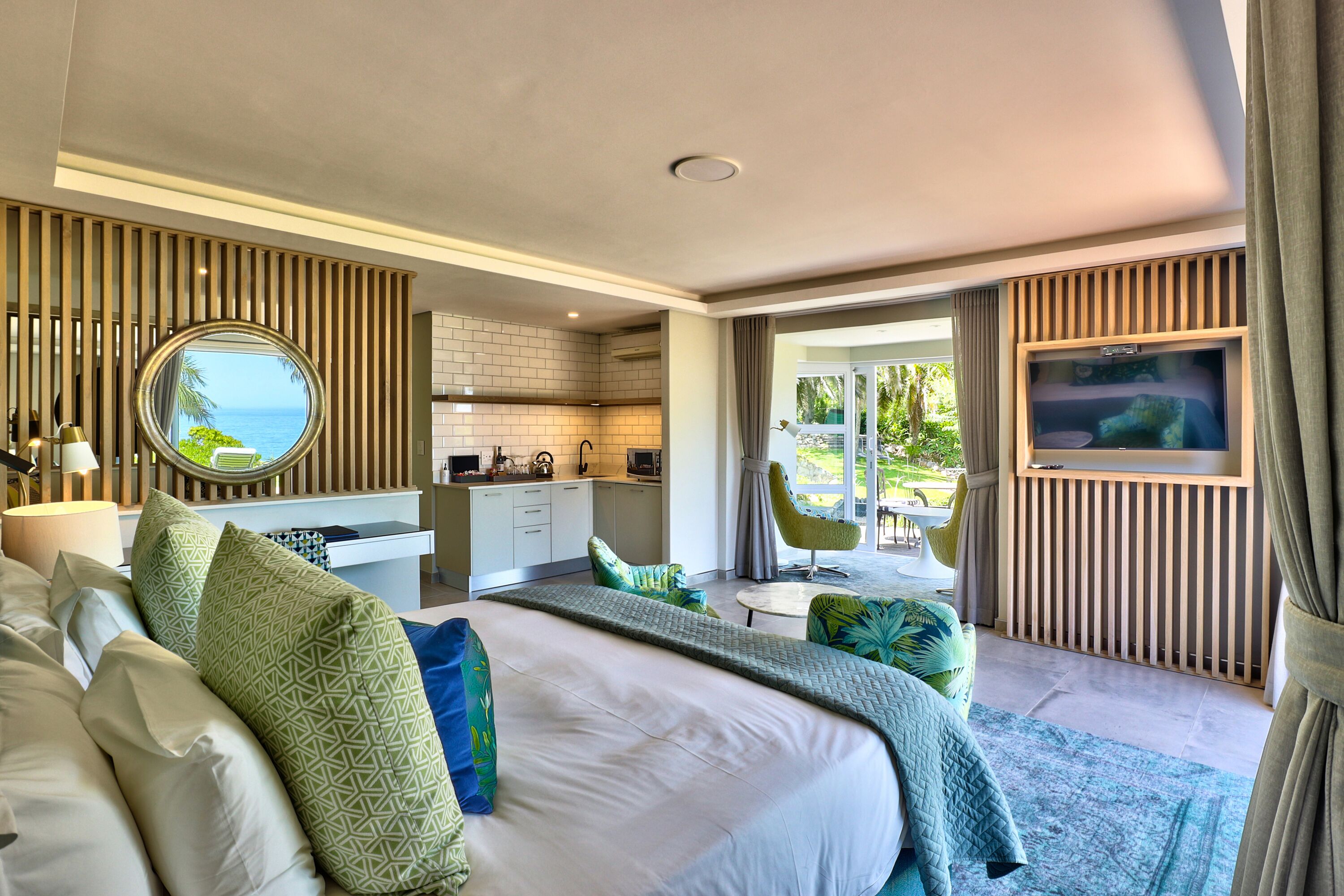 Een moderne hotelkamer met een groot bed, uitzicht op zee, een kitchenette en een balkon met een televisie.