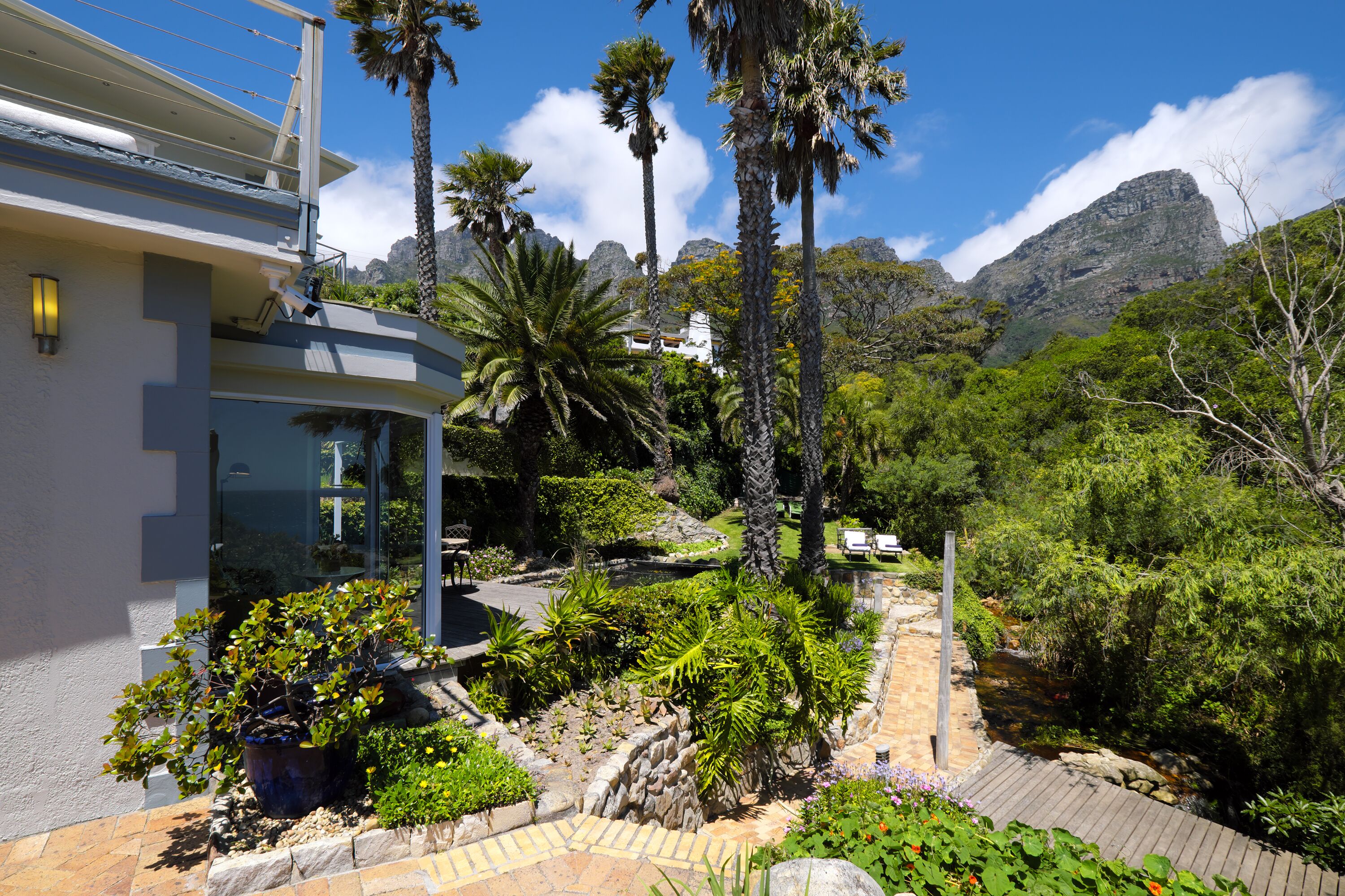 Un edificio moderno con balcón que da a un jardín ajardinado con palmeras y montañas al fondo.