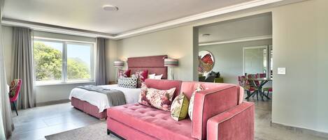 Une chambre spacieuse avec un grand lit, un canapé rose et un coin repas visible à travers une porte ouverte.