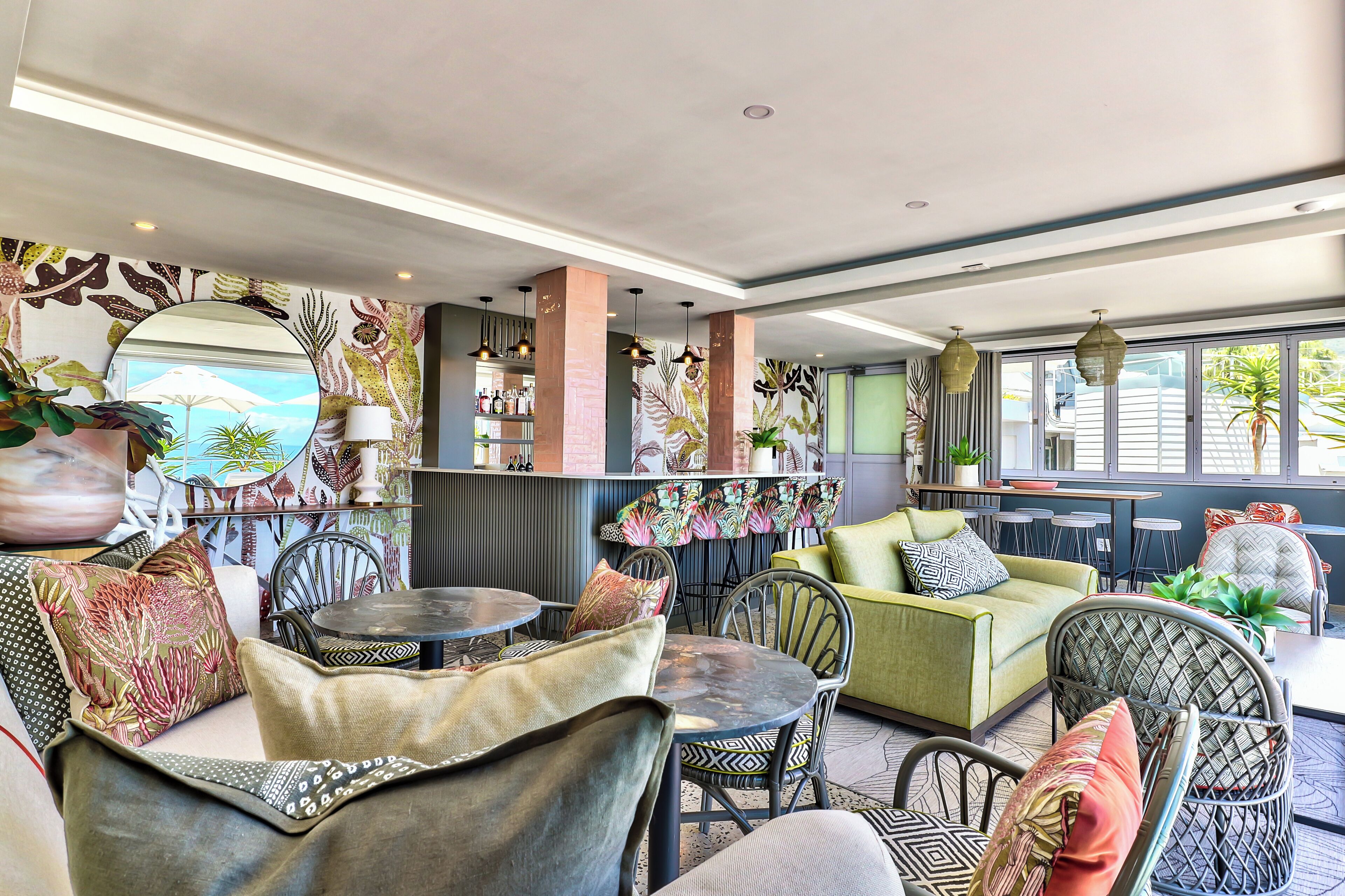 A modern lounge area with round tables, comfortable seating, a bar with a variety of bottles, and a large mirror on the wall.