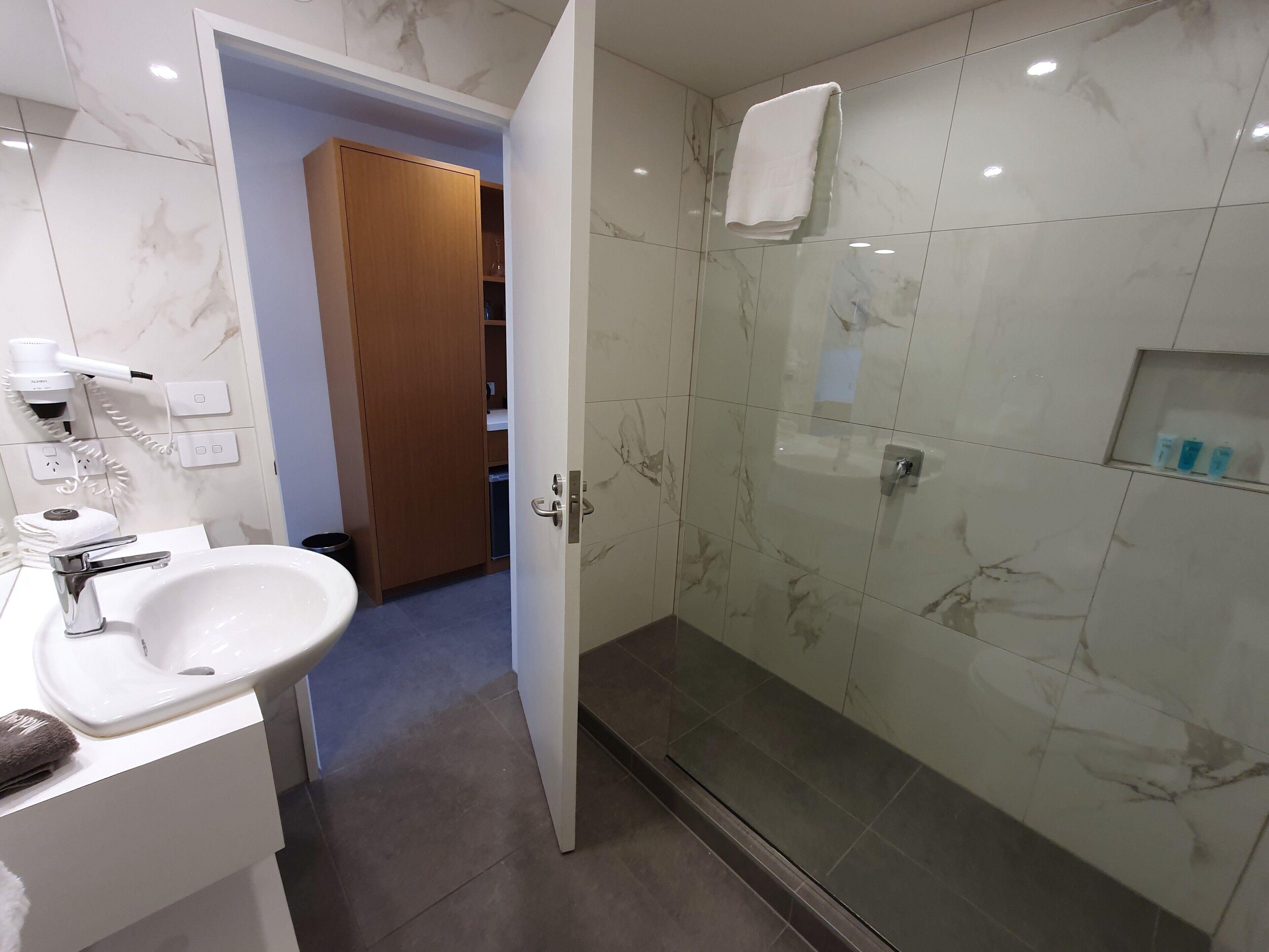 A modern bathroom with a marble-tiled shower, a white sink, and a wooden door leading to a closet.