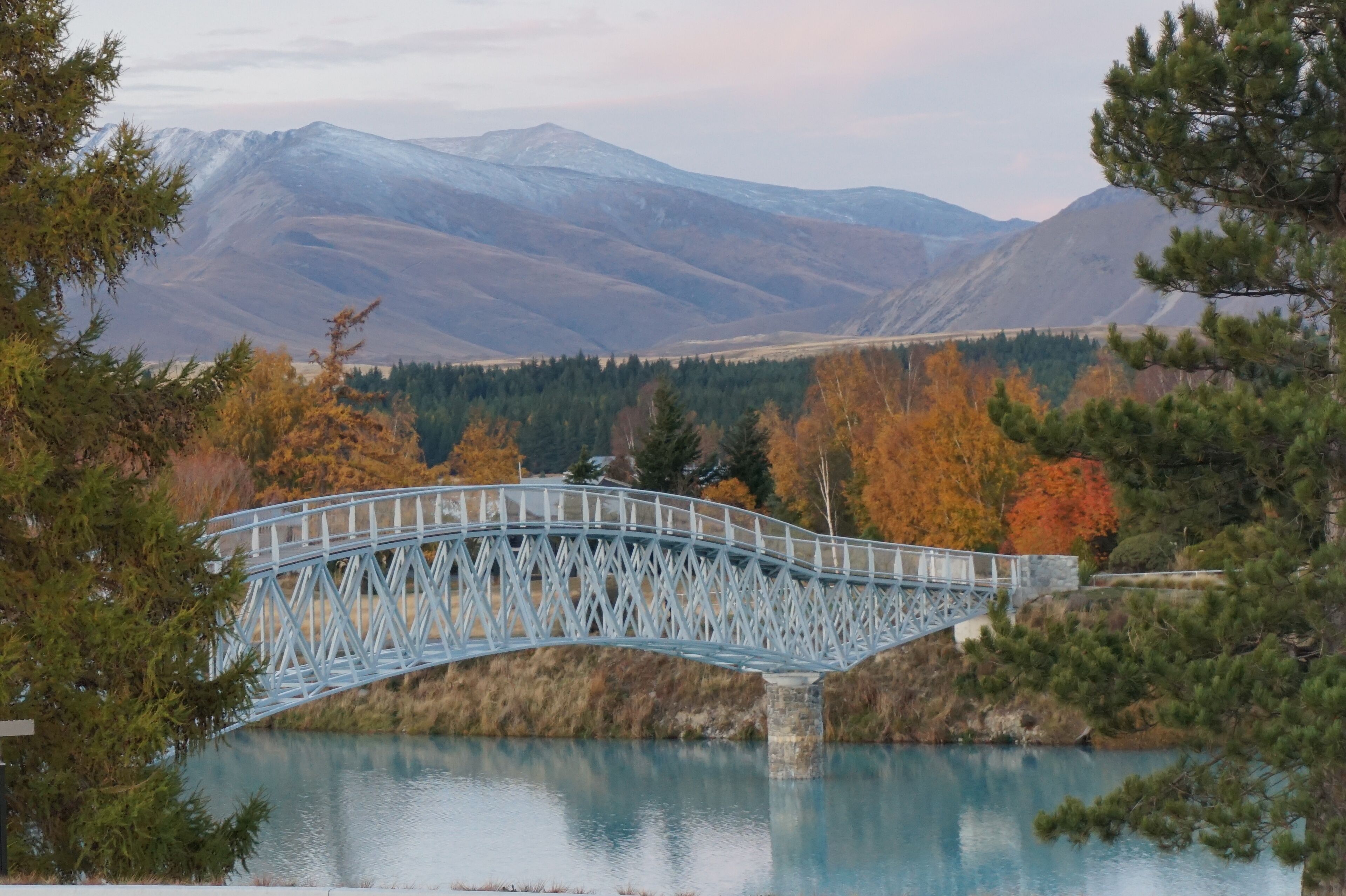 Un puente metálico sobre un lago tranquilo con montañas al fondo.