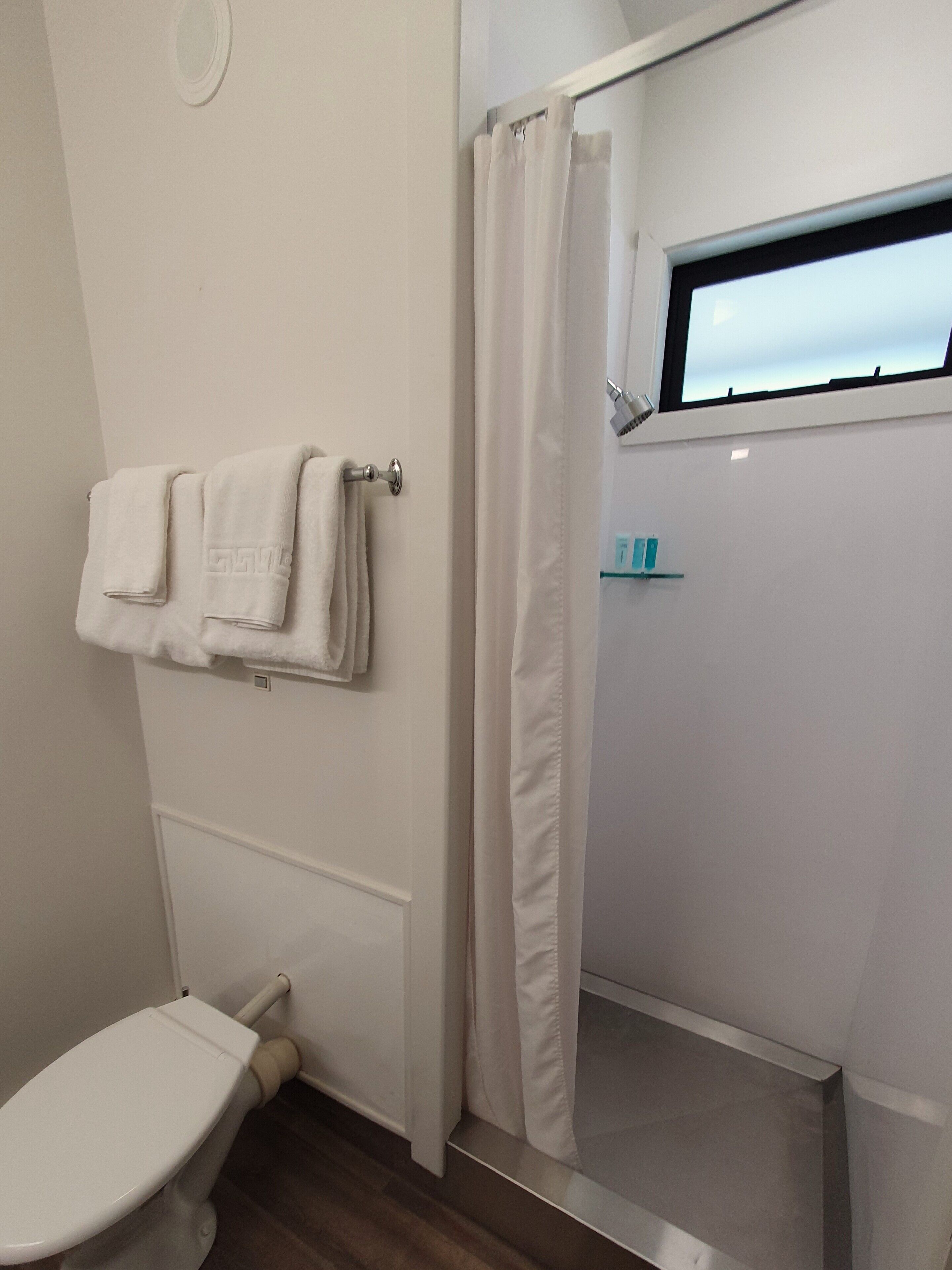A bathroom with a toilet, a towel rack with folded towels, a shower curtain, and a window.