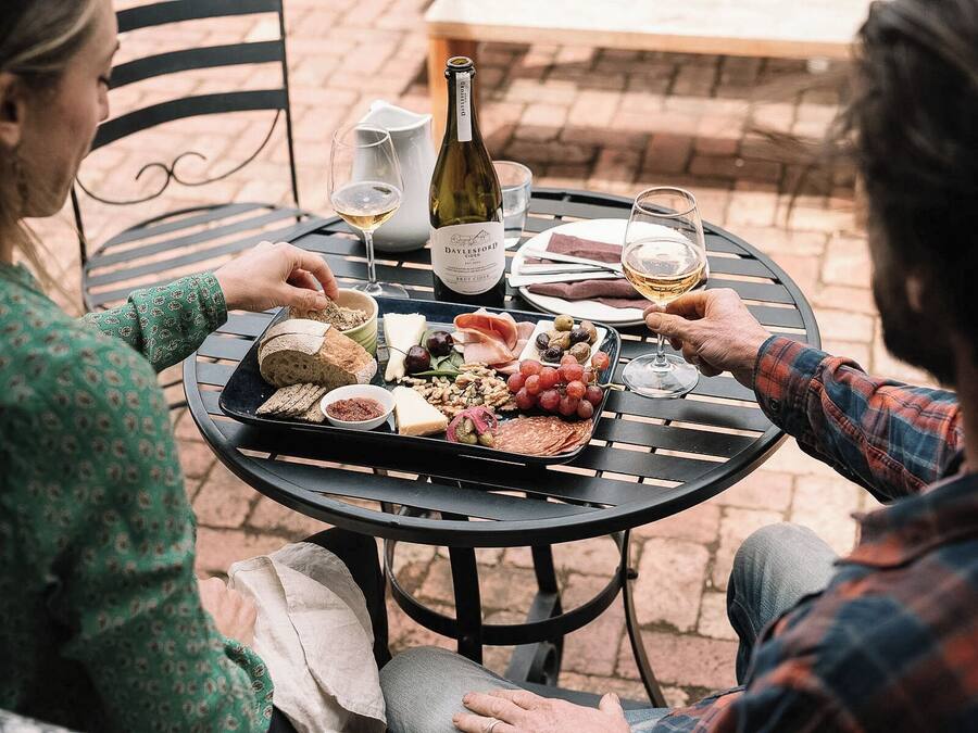Uma mesa redonda com uma garrafa de vinho, copos e uma tábua de frios com diversos queijos, carnes e frutas.