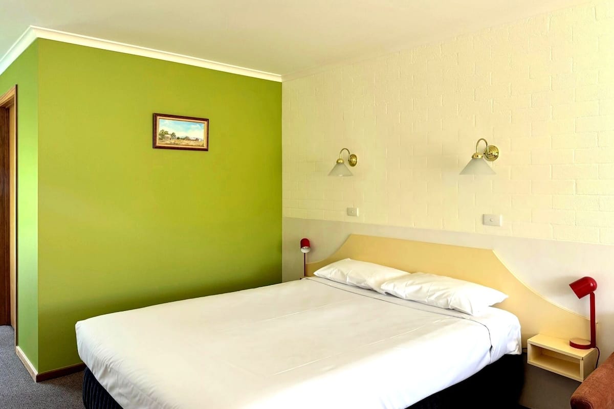 A hotel room with a bed, two bedside lamps, a nightstand, a framed picture on the wall, and a brick-patterned wall.
