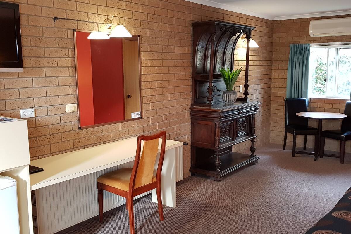 A room with a brick wall, a wooden cabinet, a desk, a chair, a table, and a window with curtains.