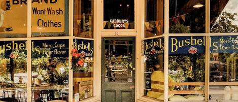 Fachada de una tienda con varios carteles que anuncian café y ropa, incluyendo 'Bushells Pure Coffee' y 'Monkey Brand Won't Wash Clothes'.