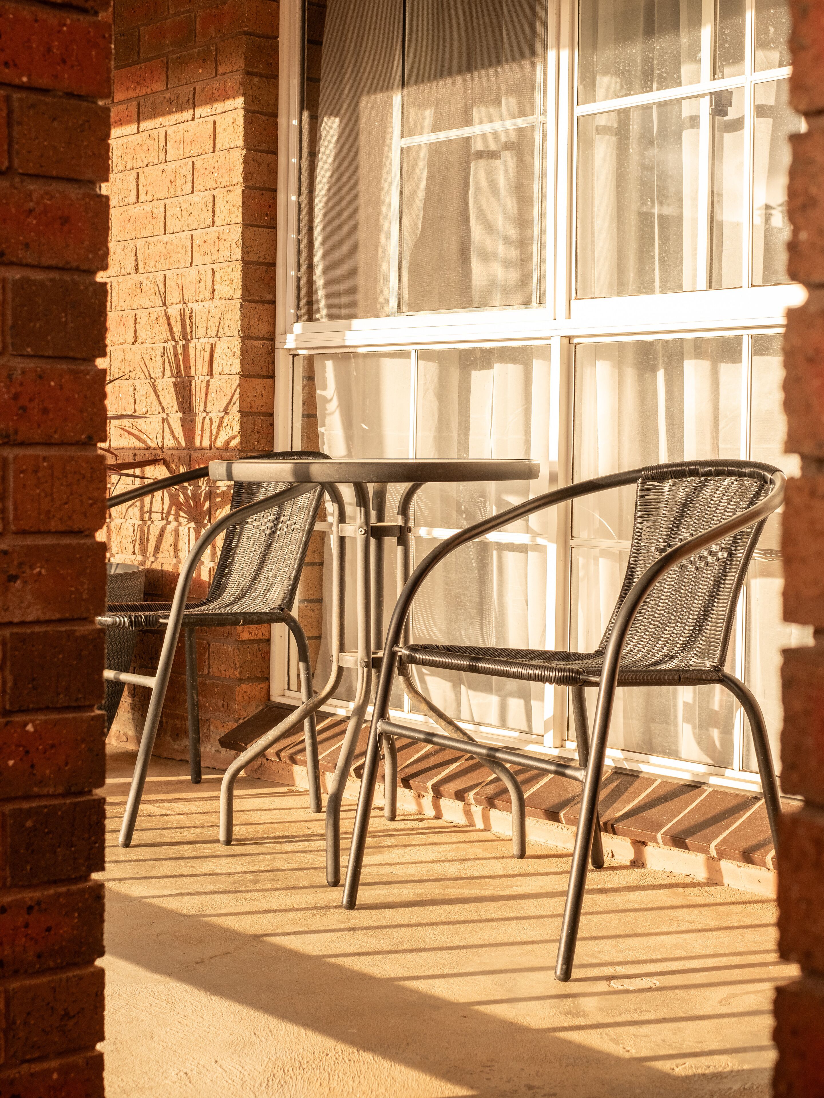 A patio with two metal chairs and a small table, a brick wall, and a window with a white curtain.