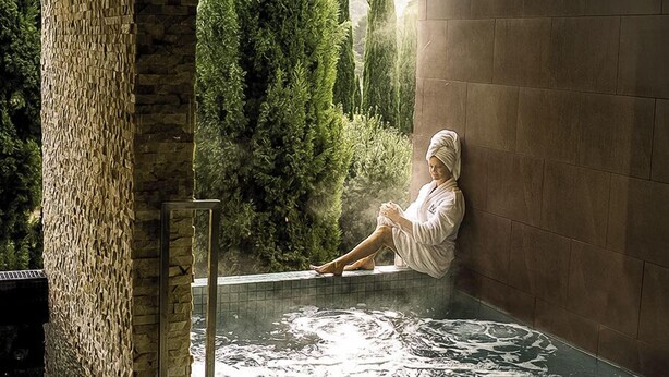 A person sitting by a stone wall in a spa area with a hot tub, surrounded by greenery.