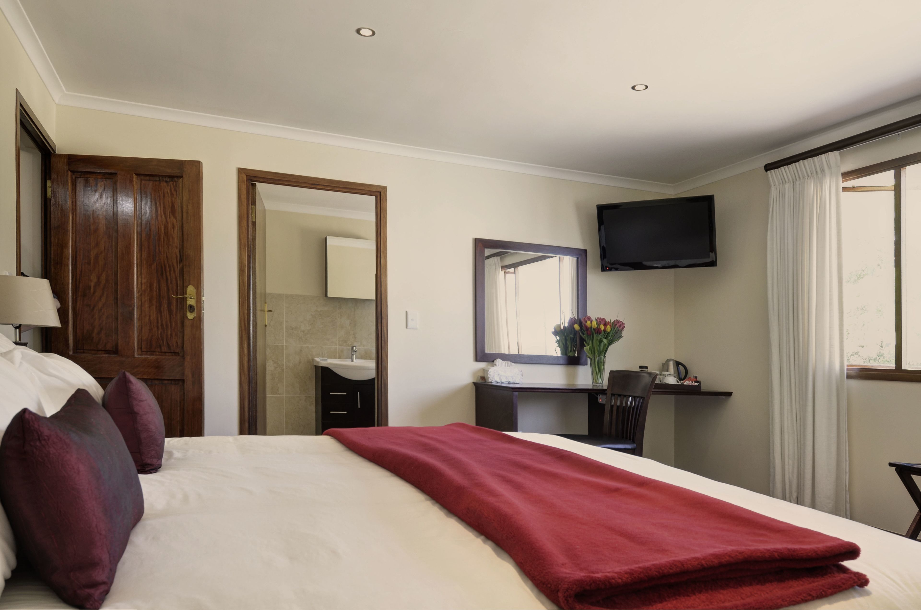 A hotel room with a bed, a TV mounted on the wall, a desk with a chair, a mirror, and a window with curtains.
