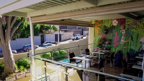 A restaurant with outdoor seating, a glass railing, and a view of parked cars and greenery. - Sundown Manor Guest House (Cape Town)