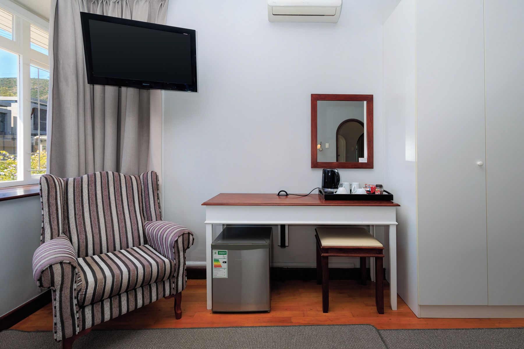 Habitación de hotel con un sillón a rayas, un escritorio con espejo, un televisor montado en la pared y un cesto de basura pequeño.
