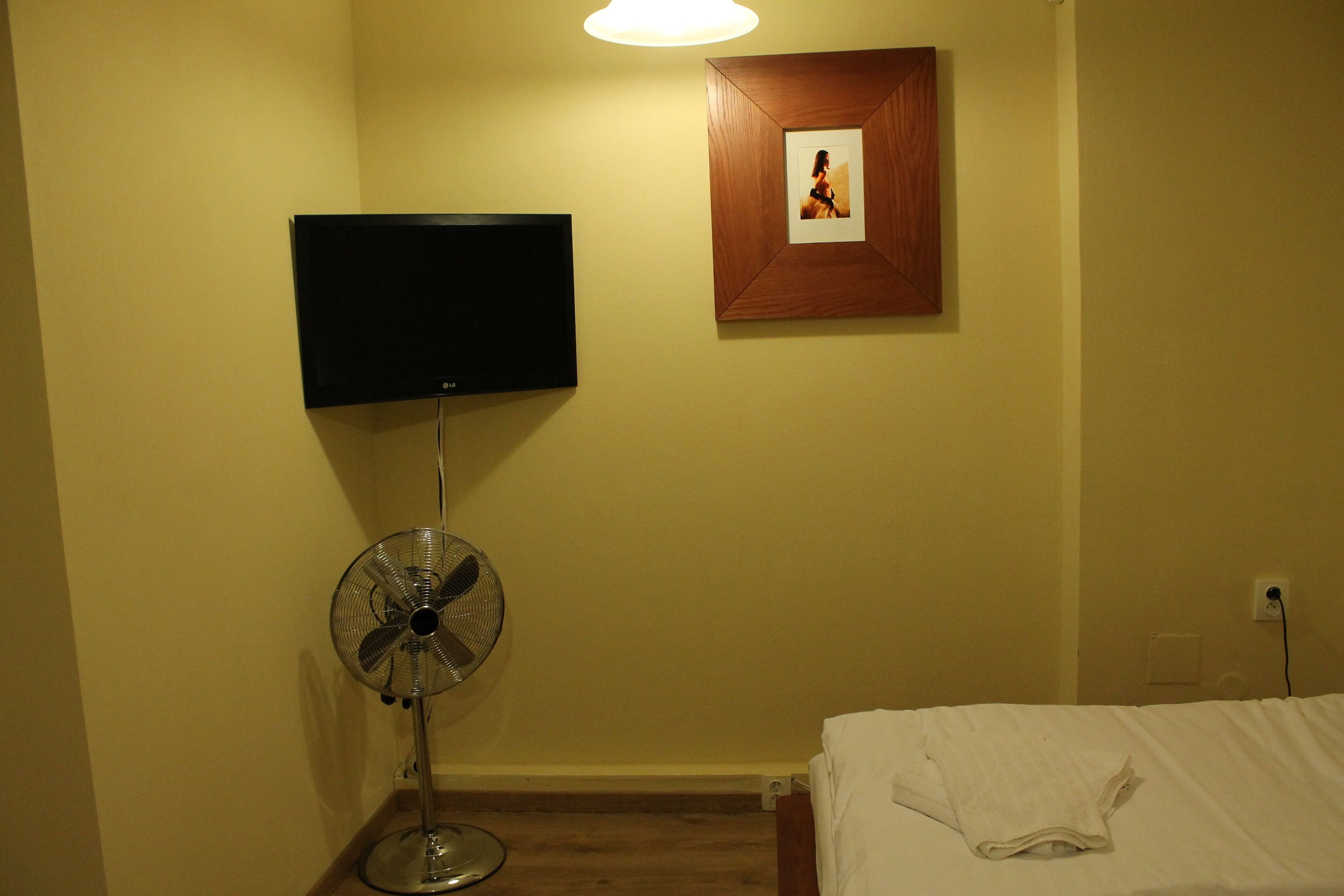 A hotel room with a bed, a standing fan, a flat-screen TV mounted on the wall, and a framed picture above the bed.