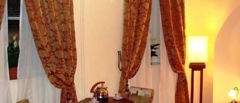 A hotel room with a bed, a small round table with a kettle, two chairs, a lamp, and curtains.