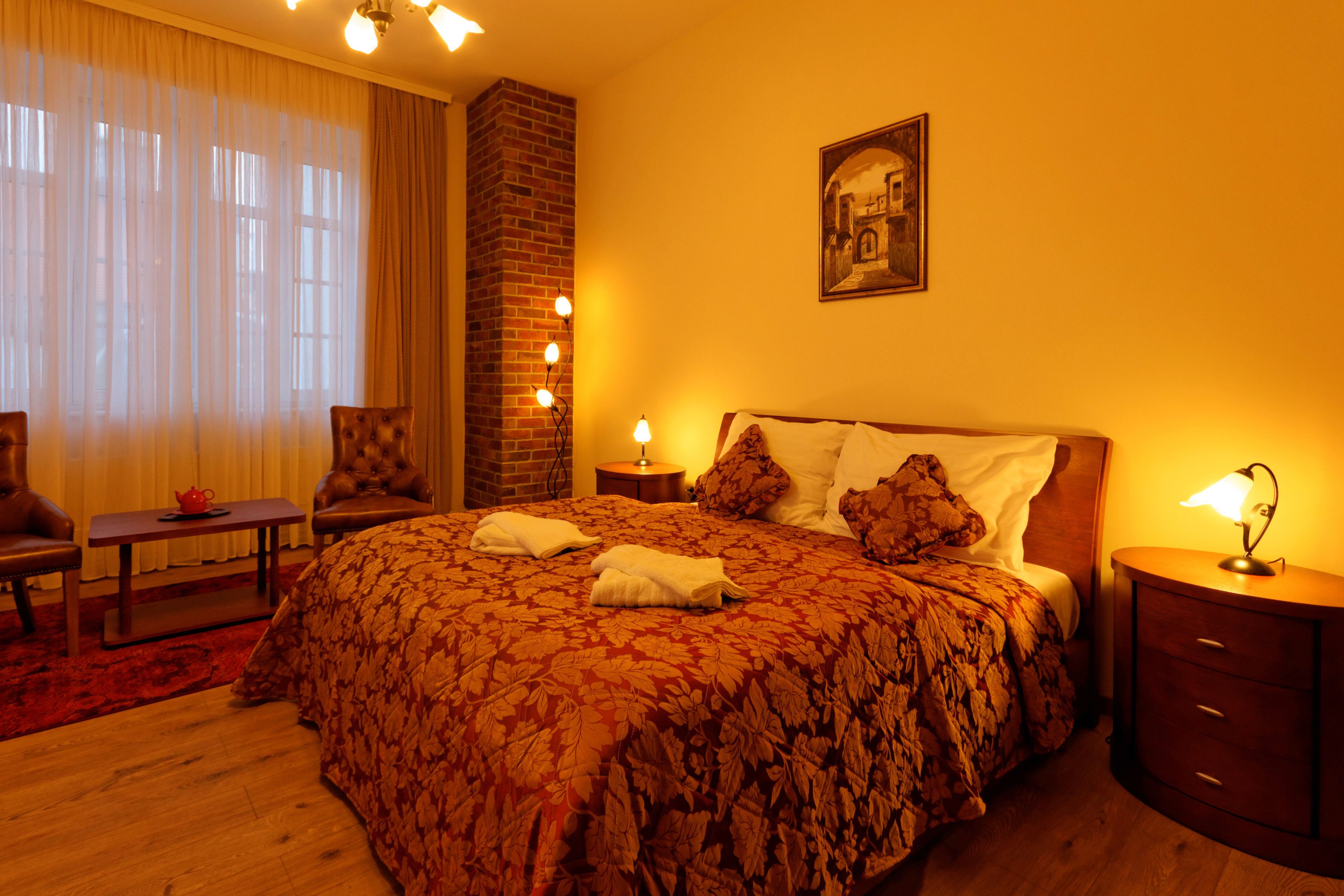 A hotel room with a large bed, a wooden nightstand, a chair, a small table, and a large window with curtains.