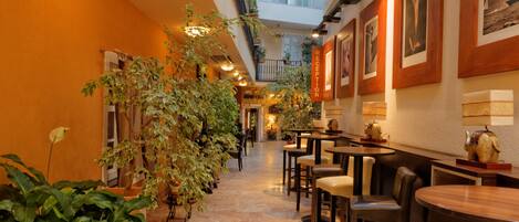 Un pasillo de hotel con techo alto, ventanas amplias y plantas de interior. Mesas y sillas están dispuestas a lo largo de las paredes.