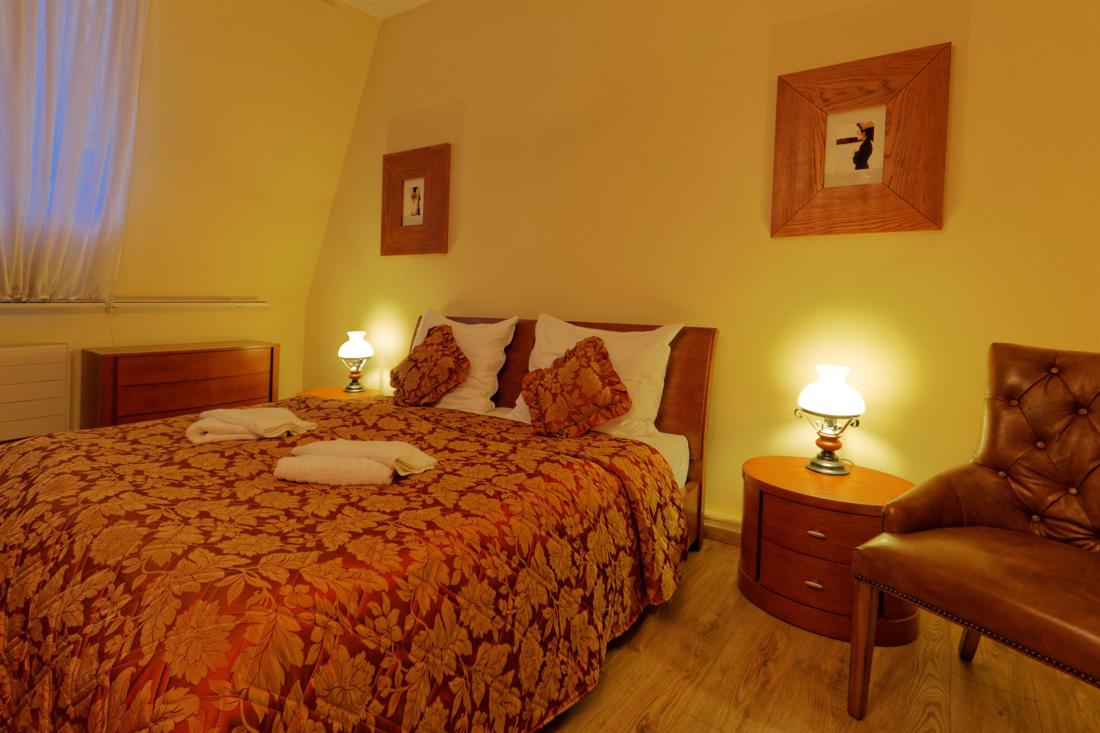 Una cama bien tendida con una colcha estampada, dos lámparas de noche, una mesita de noche de madera y un sillón de cuero.