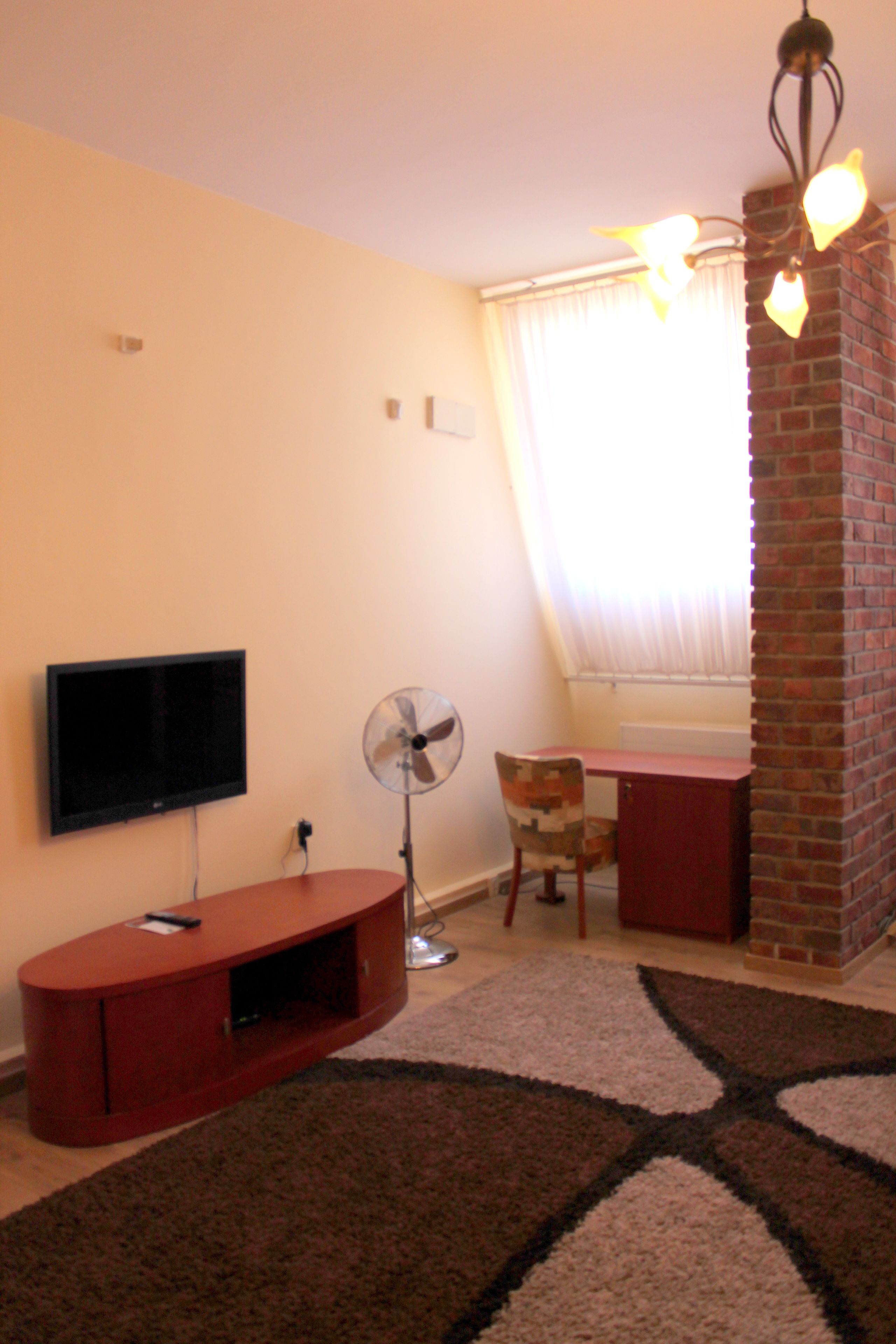 A living room with a flat-screen TV, a fan, a desk, and a brick fireplace.