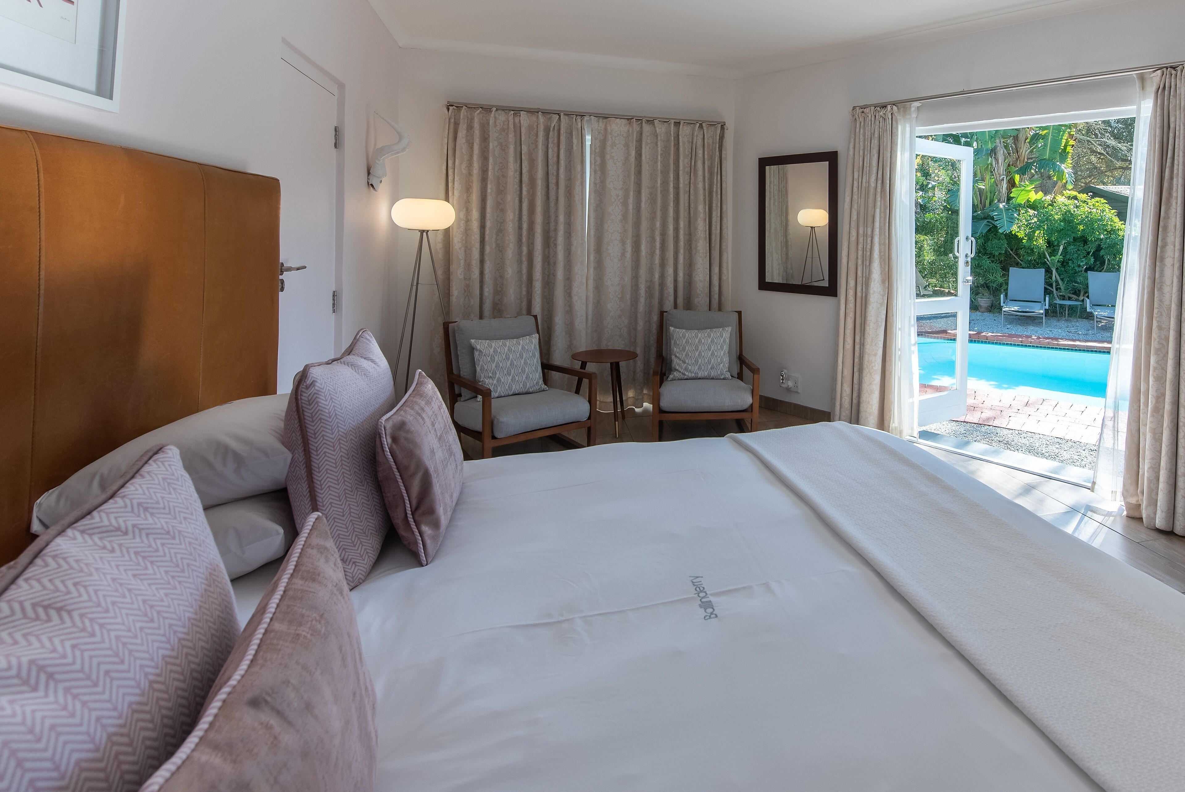 A hotel room with a large bed, two armchairs, a small table, a lamp, and a view of a swimming pool.