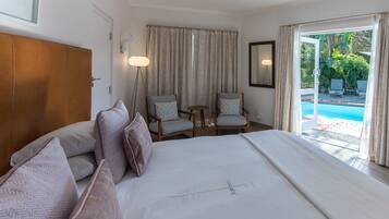 A hotel room with a large bed, two armchairs, a small table, a lamp, and a view of a swimming pool.