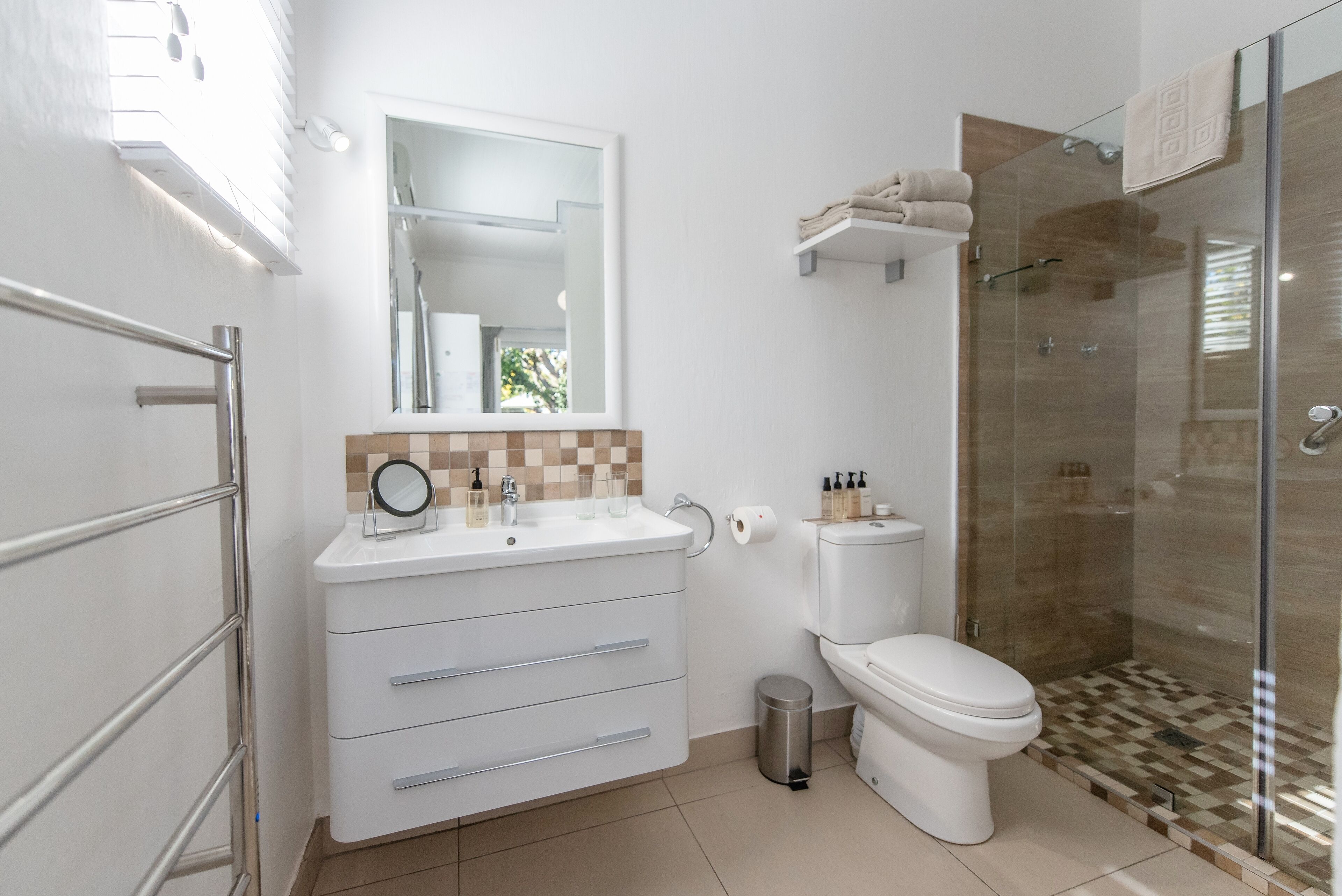 A modern bathroom with a white vanity, a toilet, a glass shower enclosure, and a small trash bin.
