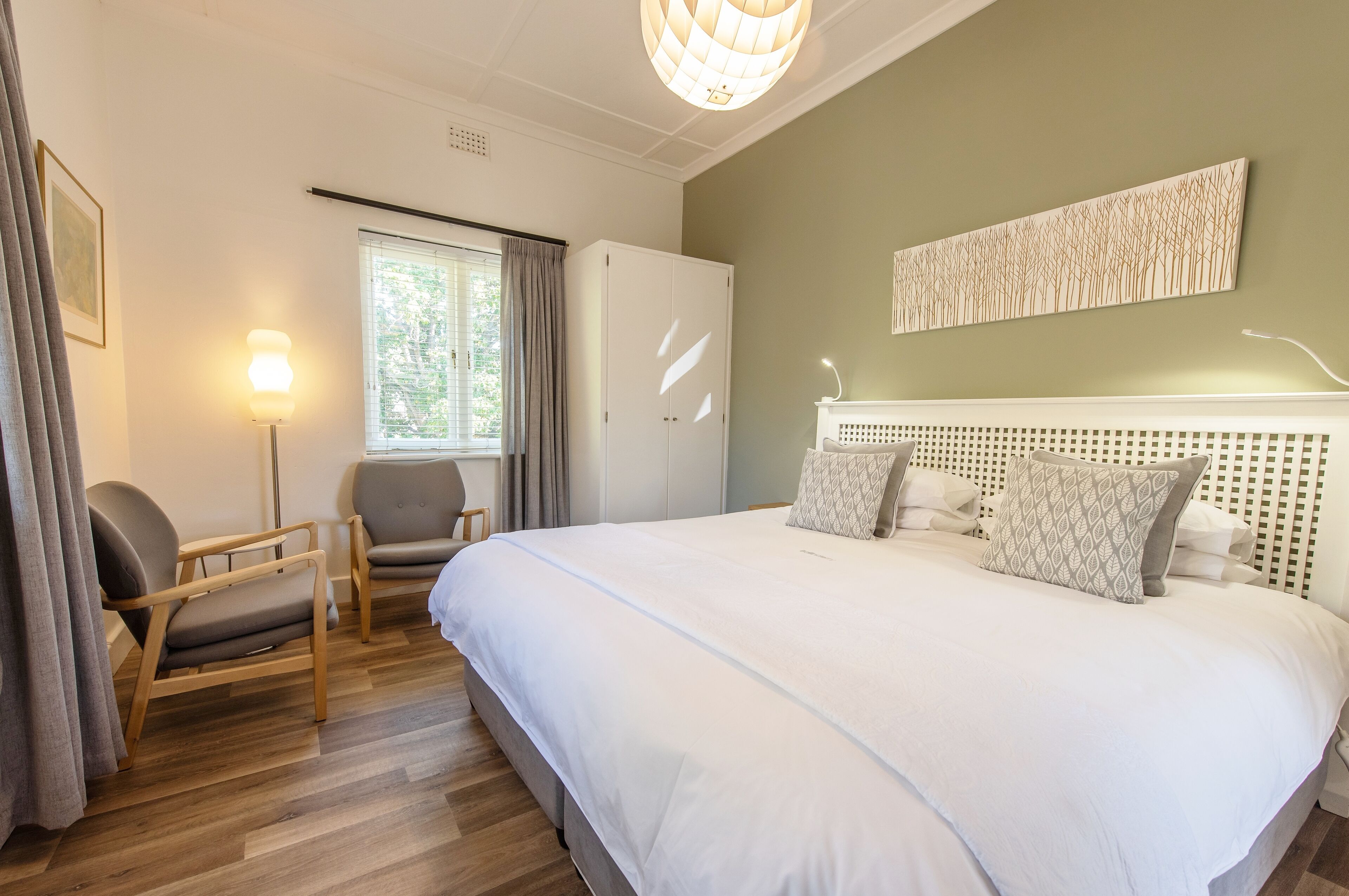 A bedroom with a large bed, two chairs, a wardrobe, a window with curtains, and a wall-mounted artwork.
