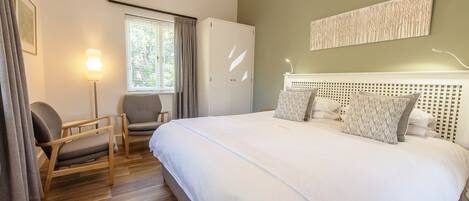 A bedroom with a large bed, two chairs, a wardrobe, a window with curtains, and a wall-mounted artwork.